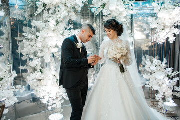 A couple stands against a backdrop of white flowers. They look at each other, and the groom puts the ring on the bride's finger.