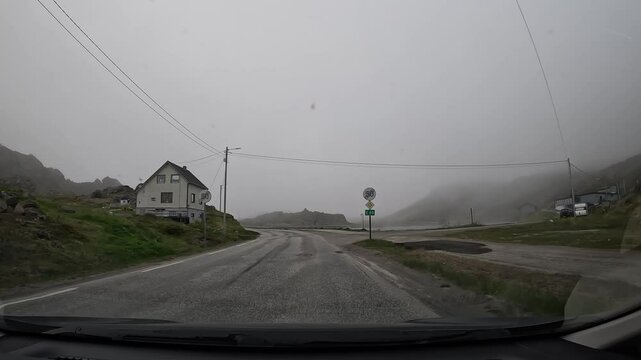 Driving through the tundra in northern Norway on E69 road. Misty scenery with rocky hills, a small lake, and few houses, creating a tranquil and remote atmosphere.