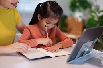 A mother is teaching her young daughter to do homework together at the table, creating a supportive and educational environment.