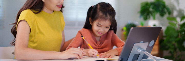A mother is teaching her young daughter to do homework together at the table, creating a supportive and educational environment.