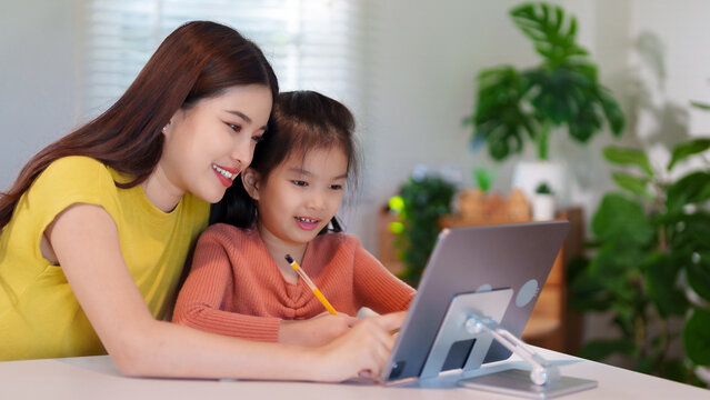 A mother is teaching her young daughter to do homework together at the table, creating a supportive and educational environment.