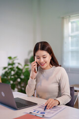A woman is multitasking, talking on the phone while using her laptop and reviewing documents on the...