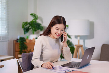 A woman is multitasking, talking on the phone while using her laptop and reviewing documents on the desk.