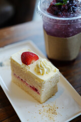 Piece of vanilla cake freshly baked strawberry pastry creamy homemade on white plated and blueberry iced soda in plastic glass on wooden table in cafe