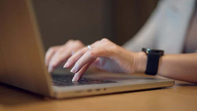 A woman is typing on a laptop computer. She is using her right hand to type and her left hand to hold the mouse. The laptop is open and the screen is lit up. The woman is focused on her work