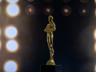 A golden Oscar statue stands on a pedestal in front of a backdrop of lights