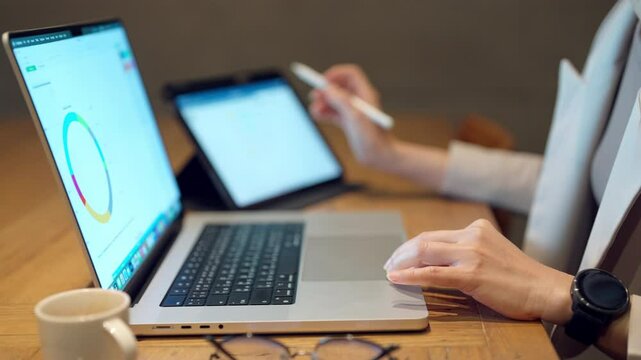 A person is using a laptop and tablet to work on a project. The laptop is open to a graph, and the tablet is open to a spreadsheet. The person is using a stylus to make changes to the spreadsheet