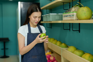 Obraz premium Young female shopkeeper smiling while holding green apple inside her fruit store. Confident woman organizing and inspecting products for quality assurance in modern retail setup.