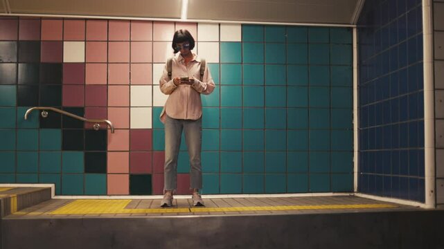 young girl is waiting for a train at the station and using the phone. viewing schedule in the application. Traveling by public transport. young girl in stylish clothes with a backpack in the subway.