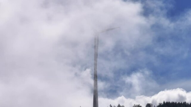 Ein Windrad dreht sich langsam vor blauem Himmel. Um das Winrad herum wandern wei&szlig;e Wolken und Nebel an einem sch&ouml;nen Sonnenaufgang. Die Sonne zeigt sich durch den vor kurzem verschwundenen Regen
