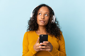 Teenager cuban girl isolated on blue background listening music with a mobile and thinking