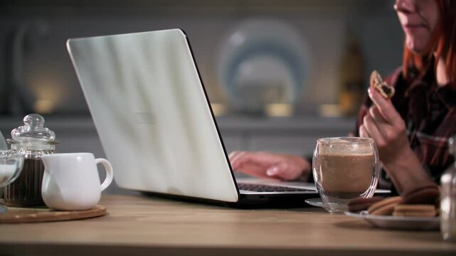 young woman eating cookies and drinking coffee sitting at kitchen table in evening at home