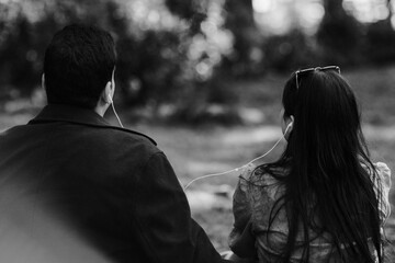A grayscale image of two individuals sitting outdoors, sharing earbuds, suggesting intimacy and connection.