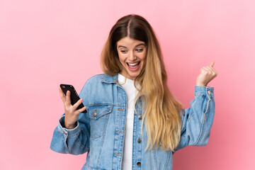 Fototapeta premium Young blonde woman isolated on blue background using mobile phone and doing victory gesture