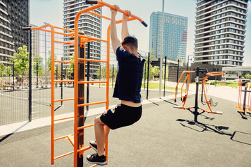 A teenager works out on the outdoor sports ground. He performs pull-ups hanging on the horizontal bar.