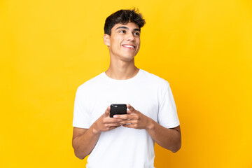 Young man over isolated yellow background using mobile phone and looking up