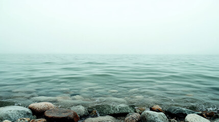 Obraz premium serene view of tranquil sea with soft green tones blending into horizon, featuring smooth rippling water and rocky shoreline in foreground under misty sky