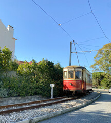 Naklejka premium Red vintage tram on a railway track surrounded by greenery and a clear blue sky.