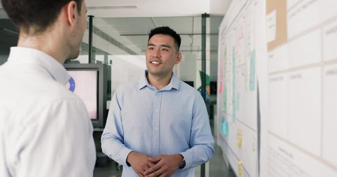 Happy Asian project manager telling scrum management strategy to business partner, investor, coworker, standing at whiteboard with sticky notes, speaking to male colleague, smiling