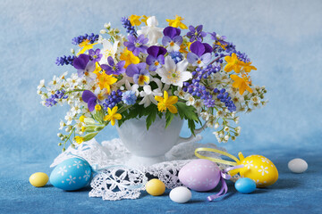 Easter beautiful card with a bouquet of spring flowers in a cup, eggs, candies on a white and blue background
