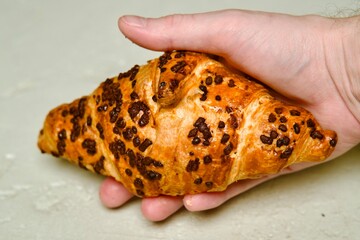 A close-up shot of a freshly baked croissant with chocolate chips, held in a hand. The image highlights the golden, flaky texture and rich chocolate topping of the pastry.