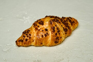 A close-up shot of a freshly baked croissant with chocolate chips, placed on a light textured surface. The image highlights the golden flaky layers and rich chocolate topping.