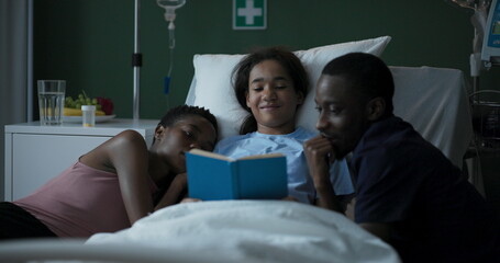 Young African girl in a hospital room with her parents sitting by the child's side reading a book together.