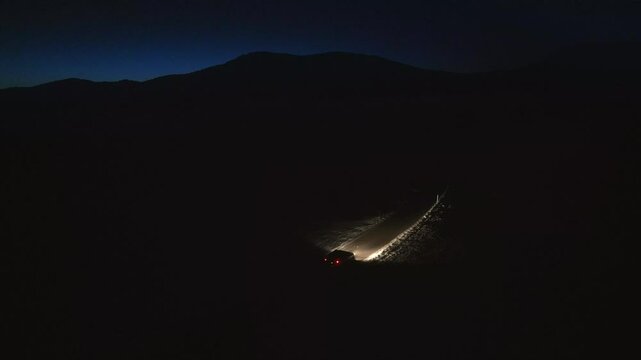 Car riding through winter forest road at night aerial tracking shot. Vehicle illuminated with headlights driving in countryside in evening drone view