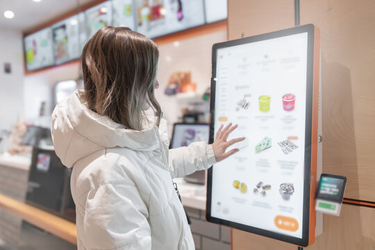 A 30 -35-year-old woman in a white winter jacket orders food through the screen menu at a fast food restaurant. A customer is utilizing a digital kiosk designed specifically for the purpose of