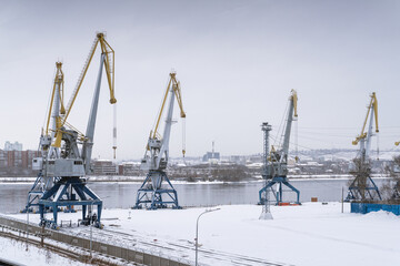 River cranes port unloading logistics at the Yenisei River in Krasnoyarsk. Cranes remain stationary in a serene winter landscape, crafting an intriguing industrial aesthetic near the river ,