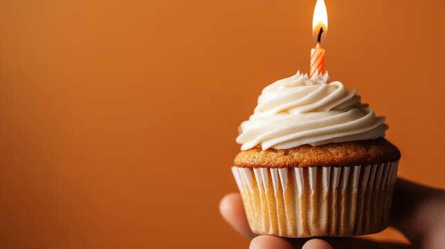 A hand presents a delectable cupcake adorned with creamy icing and a lit candle, ideal for birthdays and celebrations against a warm orange backdrop, radiating joy and festivity.