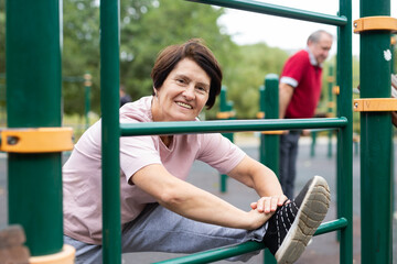 Obraz premium Positive elderly woman doing leg stretching on outdoor sports ground