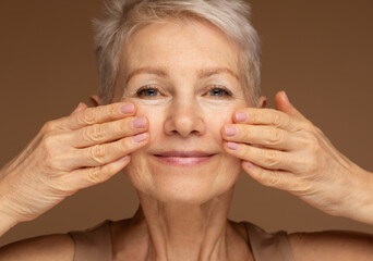 Gorgeous happy mature woman, senior older 60 year lady looking at camera touching her face over brown background.