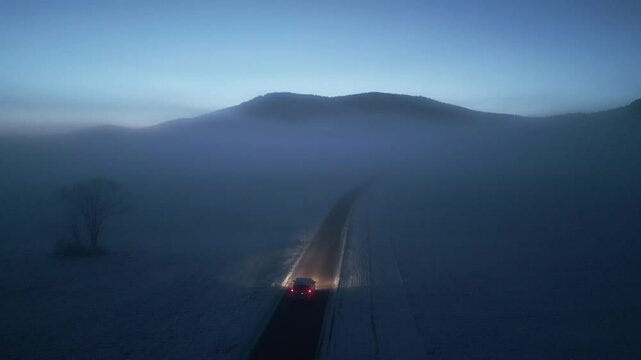 Eerie winter landscape with car on lonely road and mountains skyline aerial view. Automobile driving through morning fog on empty rural highway drone tracking shot