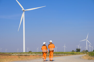 Two engineers in orange safety uniform and white helmets walking down gravel road through wind farm under clear blue sky for inspection and maintenance wind turbines. Ecology green energy industry con