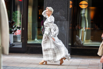 Elegant mature woman wearing long plaid dress and stylish accessories Walking confidently in urban Street near modern building, timeless fashion and sophistication. Fashion Model of Middle Age