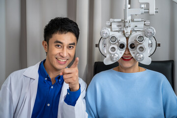 Asian male optometrist performing eye exam senior female woman with phoropter and smiling for patient care