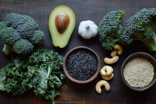 Top view of various natural, organic food items including legumes and grains on a dark wooden table, including quinoa, black rice, avocado, kale leaves, broccoli, garlic cloves, and cashew nuts.
