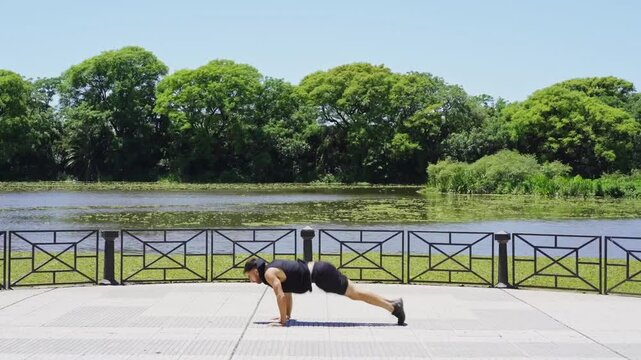 Side view of a young Latino man doing burpees by the lakeside in a city park