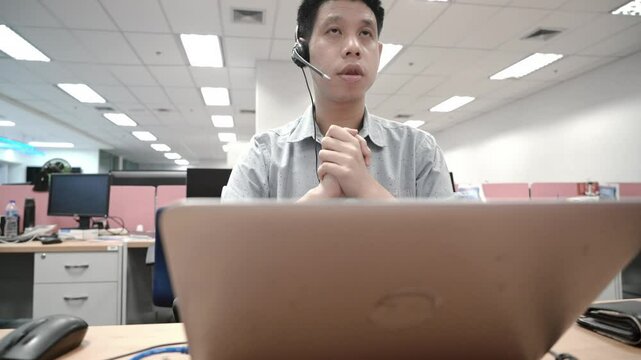 A young man in a call center demonstrates focus and concentration while wearing a headset. His hands are clasped in front of him, surrounded by a modern office environment that highlights a busy works