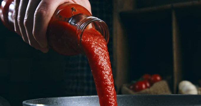 Super slow motion close up of fresh organic bio thick tomato sauce is being poured from glass jar into a frying pan for cooking pasta dish preparation in restaurant kitchen at 1000 fps.