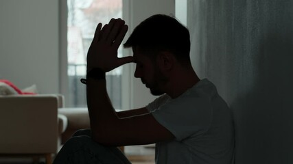 Depressed young man sitting on floor in dark room. Silhouette of young male suffering from loneliness and anxiety, covering his face with hands. Person with mental disorder - Powered by Adobe