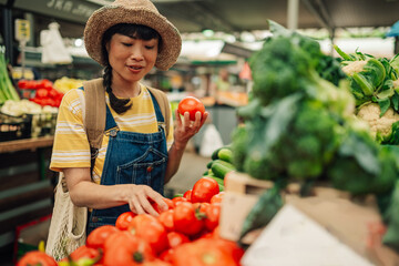 Asian woman selecting tomatoes at an outdoor farmers market stall.