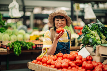 Asian woman smelling a fresh tomato at an outdoor farmers market.