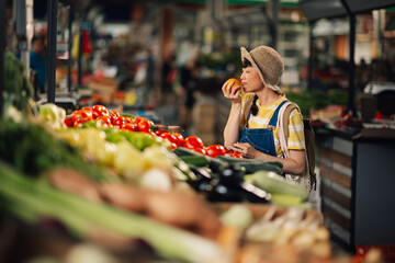 Asian woman tasting orange at market among fresh colorful vegetables