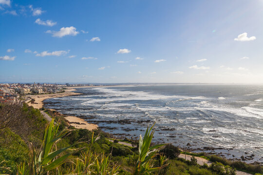 A coastal scene with a town bordering a rocky beach, waves crashing on the shore, and a blue sky with scattered clouds - Powered by Adobe