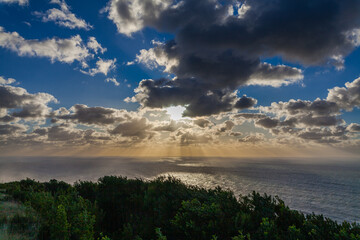 The sun breaks through dramatic clouds over a calm ocean, casting a golden light on the water.