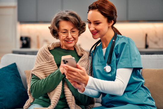 Young female doctor in blue scrubs showing an app or  medical test results or something else on her phone to an elderly woman at home. Care and recovery after illness in a medical center.