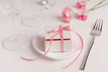 Gift box tied up with pink ribbon on ceramic plate served on white wooden table with fork, wine glasses and carnation flowers for romantic engagement holiday celebration or valentines day surprise