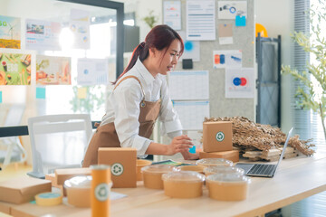 woman in apron works at desk filled with packaging materials and food containers, focused on her laptop in bright office space